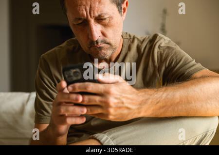 Uomo di mezza età seduto in salotto, concentrato sulla digitazione di un messaggio di testo sullo smartphone, interni casual. Messa a fuoco selettiva. Foto Stock
