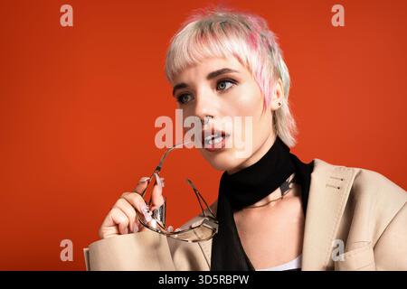 Una giovane donna sicura di sé tiene gli occhiali per le labbra, sfoggiando i suoi capelli unici e colorati. Foto Stock