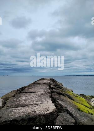 Un drammatico scatto dall'angolazione bassa che guarda giù verso un molo di pietra frastagliato che si estende nelle acque calme dell'oceano lungo una spiaggia del Connecticut Foto Stock
