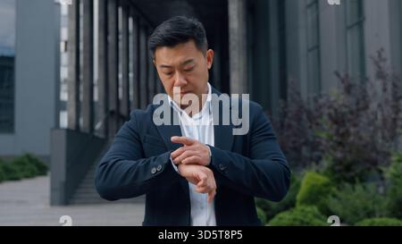 Uomo d'affari cinese di mezza età che prova ansia controllando il tempo su un orologio da polso uomo d'affari coreano asiatico in ritardo in città che aspetta affrettati a incontrare uomini aspettare Foto Stock