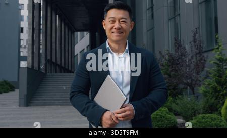 Uomo d'affari asiatico di mezza età con laptop sorridente uomo d'affari coreano che posa in città tenendo un computer sorridere felice dirigente cinese giapponese Foto Stock