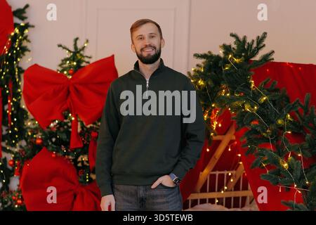 Giovane uomo sorridente con barba che posa in un casual maglione verde sullo sfondo sfocato del Natale con alberi decorati, luci e archi rossi. Vacanze fashi Foto Stock