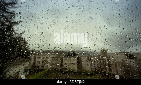 Le gocce di pioggia che scorrono giù da un'ampia finestra di casa. Primo piano, macro grandangolare, edifici sullo sfondo, nessuno Foto Stock