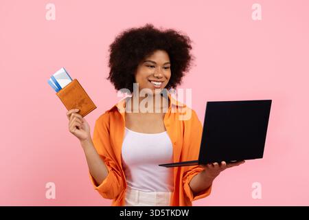 Donna afro-americana sorridente che tiene in mano passaporto e biglietti mentre usa il laptop Foto Stock