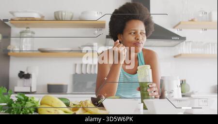 Una donna assaggiatrice in tanktop di tè assaggia il frullato verde dal vaso con cucchiaio in cucina, con frullatore Foto Stock