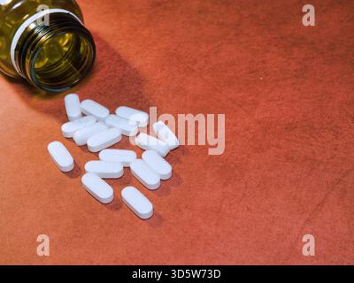 Pillole bianche fuoriuscite dal flacone - pillole oblunghe bianche fuoriuscite da un flacone di medicinali ambrato vuoto su sfondo rosso-arancione testurizzato, che simboleggia la guarigione Foto Stock