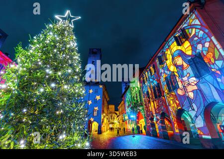 Un albero di Natale con una stella illumina una strada storica di Alba, Italia, con una proiezione luminosa colorata di un presepe sugli edifici. Foto Stock