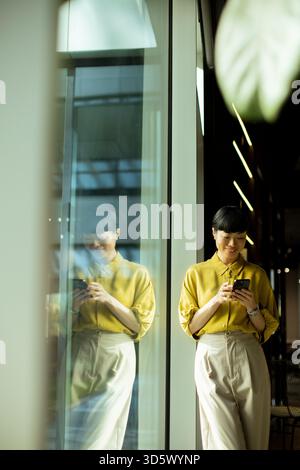 Una donna vestita con una camicia gialla e pantaloni leggeri si trova vicino a una grande finestra, guardando attentamente il suo telefono. La luce intensa filtra, creando un Foto Stock