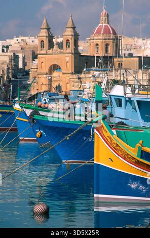 Barche da pesca colorate in un porto mediterraneo di Marsaxlock con architettura storica durante una giornata di sole. Malta Foto Stock