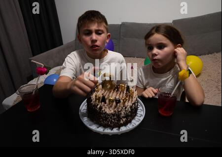 Due bambini festeggiano con entusiasmo il compleanno a casa, preparandosi a far esplodere le candele su una torta splendidamente decorata circondata da allegria Foto Stock