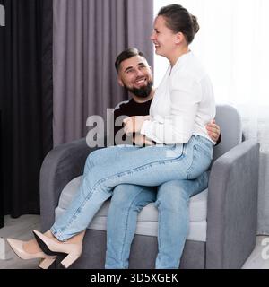 Un uomo e una donna condividono una risata gioiosa mentre si siedono insieme su un morbido divano. La luce del sole scorre attraverso la finestra, migliorando la loro connessione giocosa Foto Stock