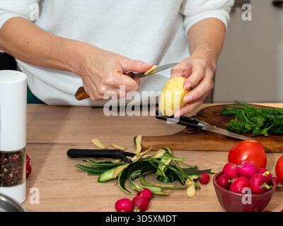 Donna che sbuccia una patata con un coltello su un bancone della cucina, circondata da verdure fresche ed erbe aromatiche. Preparazione dei pasti a casa. Foto Stock