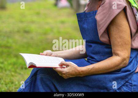 Donna anziana seduta in un parco soleggiato, che legge tranquillamente un libro e che gode di un tranquillo tempo libero all'aria aperta, immersa nel verde della natura e semplice e restaurata Foto Stock