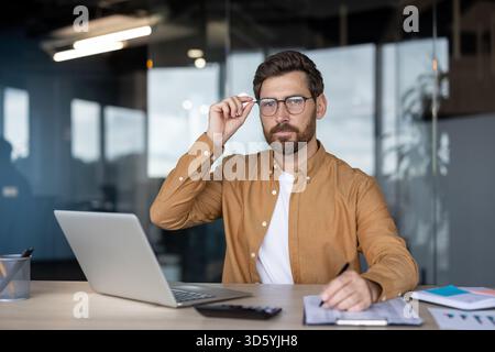 Un uomo d'affari sicuro di sé in un ufficio moderno siede alla scrivania utilizzando un computer portatile, scrivendo appunti e regolando gli occhiali mentre guarda la fotocamera, concentrato e professionale Foto Stock