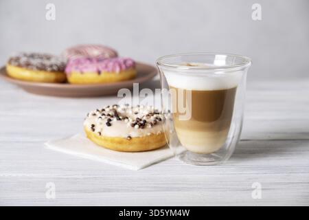 Ciambelle glassate su piatti e caffè latte o cappuccino con schiuma di latte in una tazza di vetro resistente al calore. Messa a fuoco selettiva Foto Stock