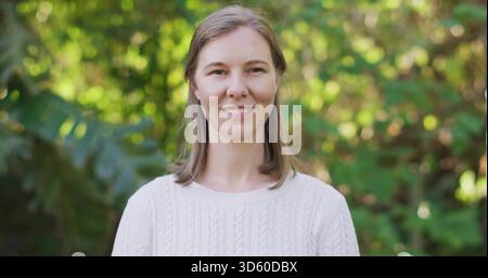 Donna in piedi in mezza età che indossa un maglione bianco in maglia di cavo e piccoli orecchini a bottone in giardino Foto Stock