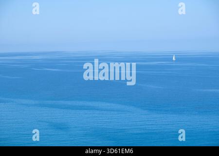 Solitaria barca a vela bianca sull'orizzonte del mare blu calmo sotto il cielo limpido della luce del giorno, minimale e serena Foto Stock