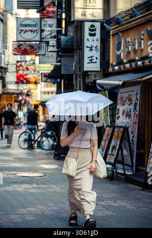 Una donna che porta un ombrellone cammina lungo un vicolo oscuro e stretto fiancheggiato da insegne tradizionali e al neon in un quartiere commerciale di Osaka, Giappone Foto Stock
