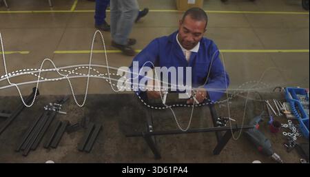 Assemblaggio di un uomo maturo con rivestimento da lavoro blu fissaggio del telaio in metallo al banco, con contenitori e trapano Foto Stock