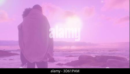 Coppia in piedi avvolta da una coperta leggera di fronte all'oceano sulla costa rocciosa, sole rosa-viola, spazio per copiare Foto Stock