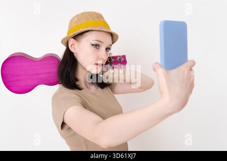 La giovane musicista si fa selfie con il telefono cellulare, mentre l'ukulele le si aggrappa sulla spalla. Elegante donna caucasica di 18 anni ukulelist posa con h Foto Stock