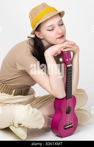 Giovane musicista femminile rilassata con gli occhi chiusi, che poggia il mento sulle mani, appoggiato su un ukulele verticale, seduta a croce con le gambe sul pavimento. Donna musi Foto Stock