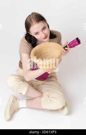 Musicista donna con vista ad alto angolo con ukulele che regge il cappello di paglia davanti, pronta a richiedere contributi finanziari, mance. Lei si sedeva a gambe incrociate Foto Stock