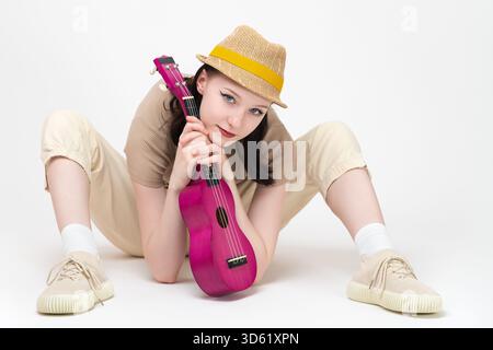 Una musicista con l'ukulele tra le mani si siede sul pavimento con le gambe separate e uno strumento musicale tra loro, appoggiandosi su di esso e guardando la telecamera W. Foto Stock
