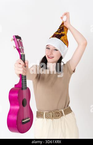 Felice e sorridente giovane musicista che indossa il cappello di Babbo Natale tiene l'ukulele davanti a una mano e tiene il cappello di pompon con l'altra, sorride giocosamente e frizzante Foto Stock