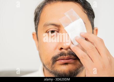 Primo piano, un uomo triste di Asain che copre il suo occhio con la benda su uno sfondo bianco. Concetto di dolore agli occhi. Foto Stock