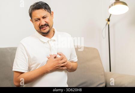 Uomo asiatico maturo seduto sul divano stringendosi e avendo dolore al petto causa di attacco di cuore. Concetto di assicurazione sanitaria per persone mature. Foto Stock