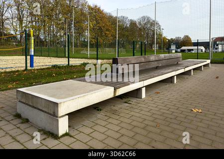 Lunghe panche in cemento e legno fiancheggiano un'area sportiva con un campo da pallavolo e da calcio visibili. Le foglie autunnali e gli alberi sullo sfondo creano Foto Stock
