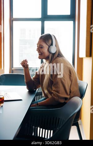 Una giovane donna allegra che indossa le cuffie partecipa a una riunione virtuale presso una scrivania, trasmettendo un senso di comunicazione, produttività e positivo Foto Stock