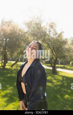 Una giovane donna sorride felice mentre ascolta musica con le cuffie, durante una passeggiata nel parco, Saragozza, Spagna Foto Stock