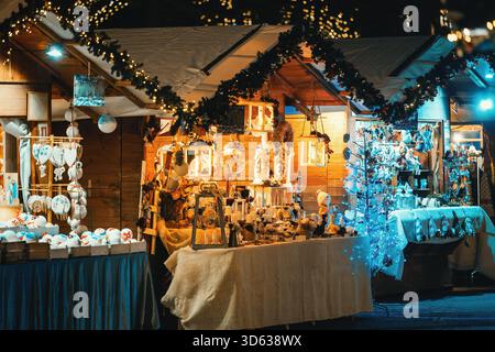Decorazioni e oggetti d'artigianato festivi sono esposti in vendita su bancarelle in legno decorate, illuminate di notte al mercatino di Natale di Asti, Italia. Foto Stock