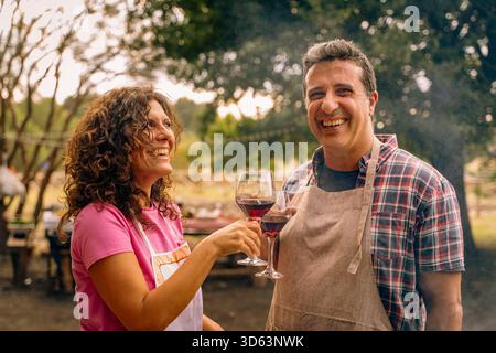Due persone che indossano grembiuli sorridono e brindano con bicchieri da vino in un allegro raduno all'aperto con alberi sullo sfondo. Foto Stock