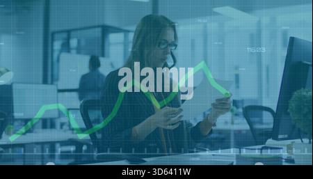 Revisione di documenti, donna in blazer e occhiali che studiano sovrapposizione di grafici con monitor in ufficio aperto Foto Stock