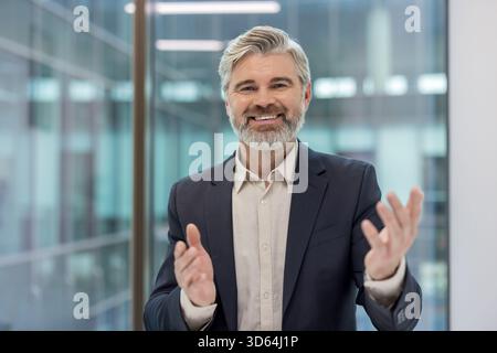 Uomo d'affari maturo e sicuro con barba grigia e tuta che applaudono e sorridono in un ufficio moderno, celebrando i risultati raggiunti e mostrando approvazione e soddisfazione professionale Foto Stock