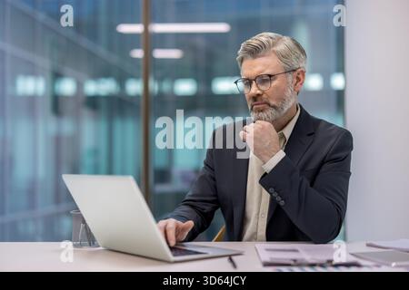 Uomo d'affari esperto che lavora su un notebook in un ufficio moderno, pianifica una strategia aziendale concentrandosi su un'attività impegnativa, creando un ambiente professionale e mirato Foto Stock