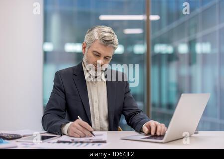 Un uomo d'affari maturo, in tuta, studia i rapporti finanziari e lavora su un laptop alla scrivania dell'ufficio, concentrandosi su grafici, grafici e dati mentre pianifica la strategia in un ambiente aziendale moderno Foto Stock