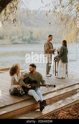 Un gruppo di amici si rilassa in riva al lago, condividendo risate e musica mentre si gusta un drink rinfrescante in un pomeriggio tranquillo. Foto Stock