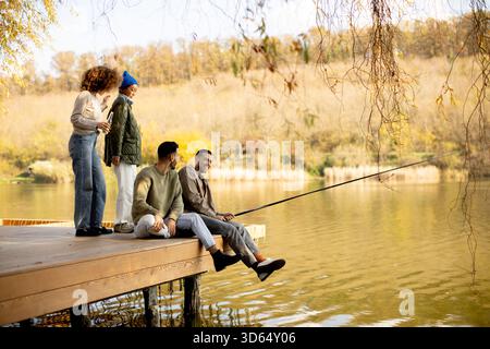 Quattro amici si riuniscono su un molo di legno vicino a un lago tranquillo, godendosi la pesca in una giornata d'autunno soleggiata. Risate e cameratismo riempiono l'aria mentre condividono Foto Stock