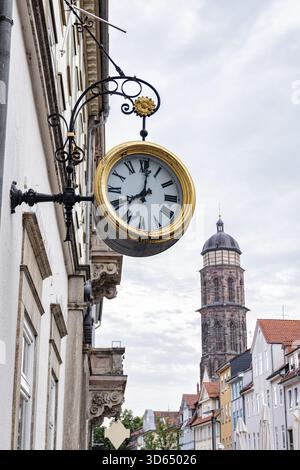 Orologio da strada d'epoca con numeri romani appesi su un edificio storico, con la torre della chiesa di San Giovanni sullo sfondo a Göttingen, Germania Foto Stock