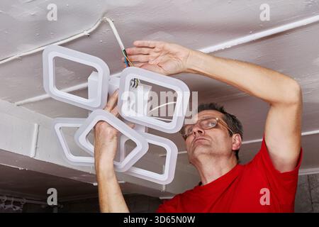 L'elettricista dell'impianto solleva il nuovo lampadario a LED verso il soffitto, il cablaggio esposto è visibile, il dispositivo è allineato al punto di montaggio, lavori in corso. Foto Stock