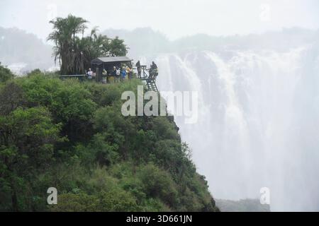 Blick auf einen Aussichtspunkt im Park Victoria Falls auf die Victoria Fälle, Die sich im Hintergrund befinden. während die linke Seite zu Simbabwe gehört, ist die rechte seite Staatsgebiet von Sambia. BEI den Victoriafällen donnert das Wasser des Sambesi auf einer Breite von 1,700 m Breite 110 m tief in eine 50 m Breite Schlucht. In der Regenzeit cadde hier bis zu 5,000 Kubikmeter Wasser pro minuto in Die Schlucht. DAS foto ist von der Victoria Fall Brücke aufgenommen, Die Simbabwe mit Sambia verbindet. Auf der linken Seite liegt Simbabwe und auf der rechten Sambia. Länge des Sambesi: 2,660 Foto Stock