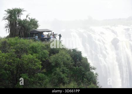 Blick auf einen Aussichtspunkt im Park Victoria Falls auf die Victoria Fälle, Die sich im Hintergrund befinden. während die linke Seite zu Simbabwe gehört, ist die rechte seite Staatsgebiet von Sambia. BEI den Victoriafällen donnert das Wasser des Sambesi auf einer Breite von 1,700 m Breite 110 m tief in eine 50 m Breite Schlucht. In der Regenzeit cadde hier bis zu 5,000 Kubikmeter Wasser pro minuto in Die Schlucht. DAS foto ist von der Victoria Fall Brücke aufgenommen, Die Simbabwe mit Sambia verbindet. Auf der linken Seite liegt Simbabwe und auf der rechten Sambia. Länge des Sambesi: 2,660 Foto Stock