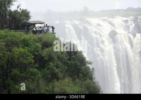 Blick auf einen Aussichtspunkt im Park Victoria Falls auf die Victoria Fälle, Die sich im Hintergrund befinden. während die linke Seite zu Simbabwe gehört, ist die rechte seite Staatsgebiet von Sambia. BEI den Victoriafällen donnert das Wasser des Sambesi auf einer Breite von 1,700 m Breite 110 m tief in eine 50 m Breite Schlucht. In der Regenzeit cadde hier bis zu 5,000 Kubikmeter Wasser pro minuto in Die Schlucht. DAS foto ist von der Victoria Fall Brücke aufgenommen, Die Simbabwe mit Sambia verbindet. Auf der linken Seite liegt Simbabwe und auf der rechten Sambia. Länge des Sambesi: 2,660 Foto Stock