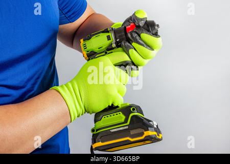 Foto di un lavoratore maschio che indossa guanti verdi protettivi che tengono un avvitatore senza fili su sfondo sgranato. Foto Stock