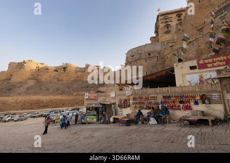 Venditori e piccoli negozi alla base del forte di Jaisalmer, con bastioni di arenaria dorata e attività serali intorno al monumento storico. Foto Stock