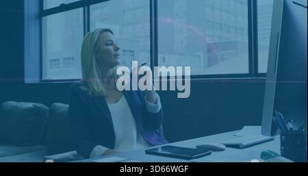 Con la penna, la donna con il blazer blu navy, la camicetta bianca che guarda le grandi finestre dell'ufficio con un tablet Foto Stock